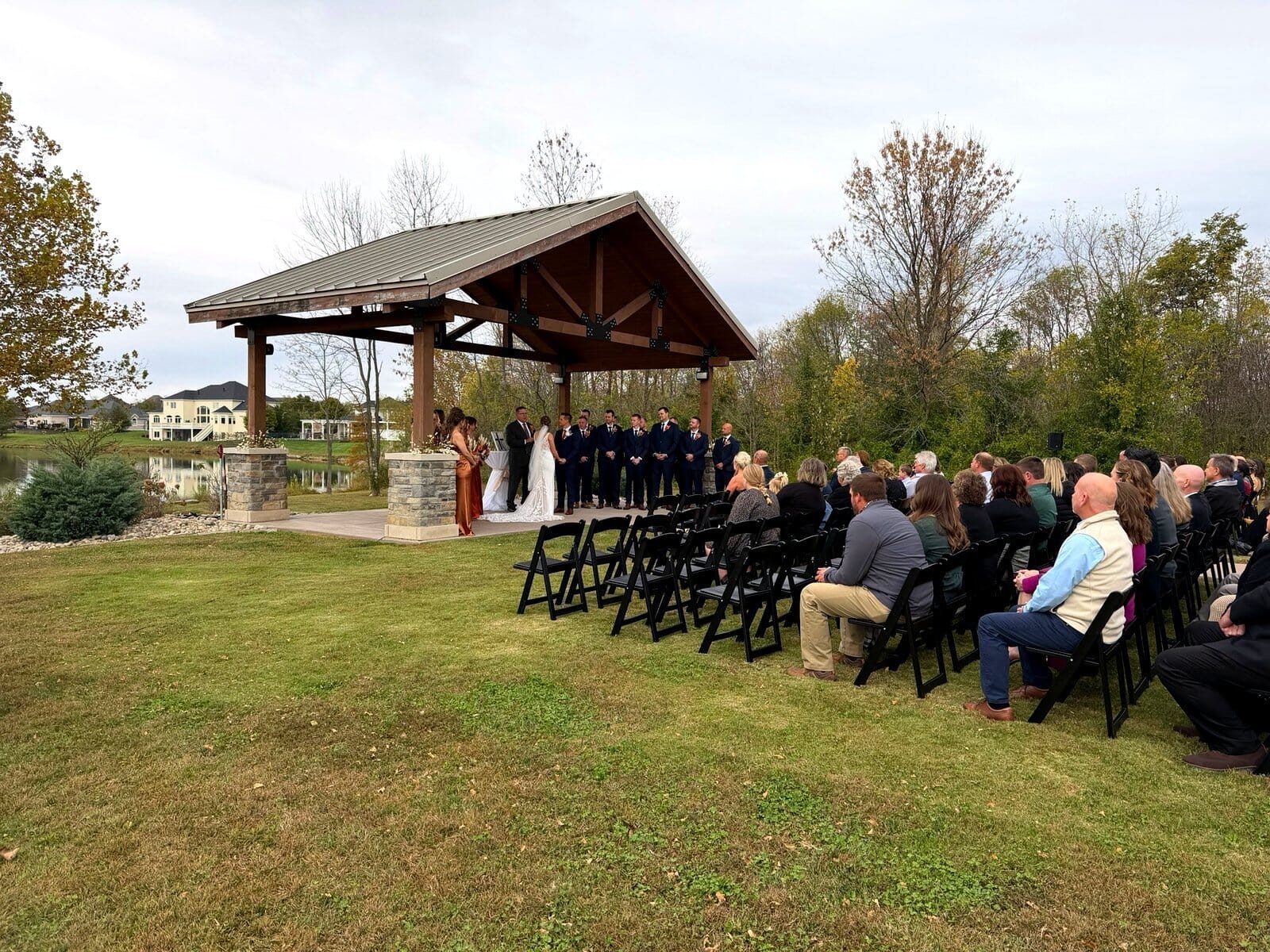Outdoor wedding ceremony with a bride and groom under a wooden pavilion at Friedman Park in Newburgh, surrounded by their wedding party and guests.
