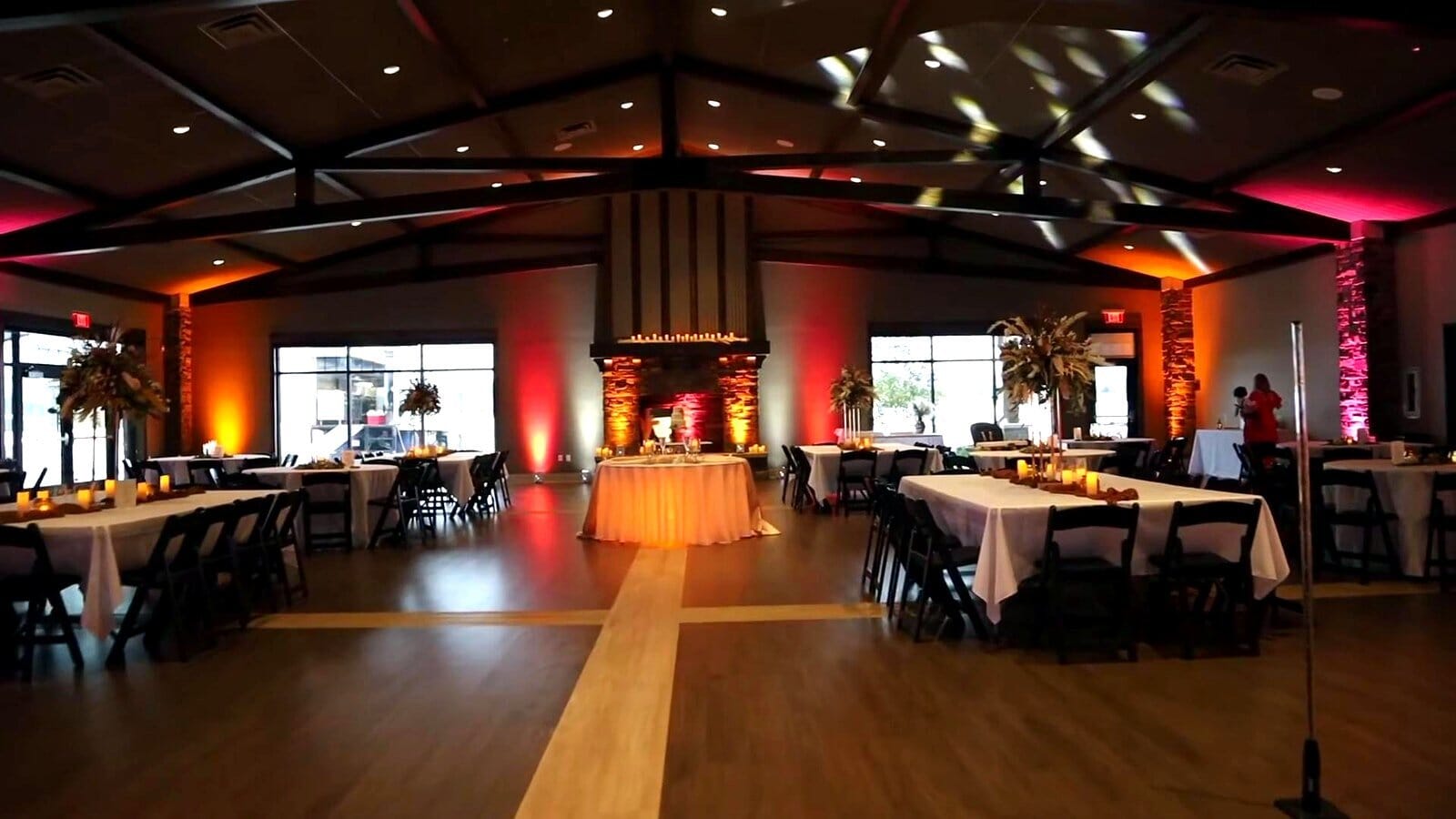 Friedman Park Event Center reception room with exposed wood beams, stone fireplace wall and warm uplighting, tables set around the dance floor