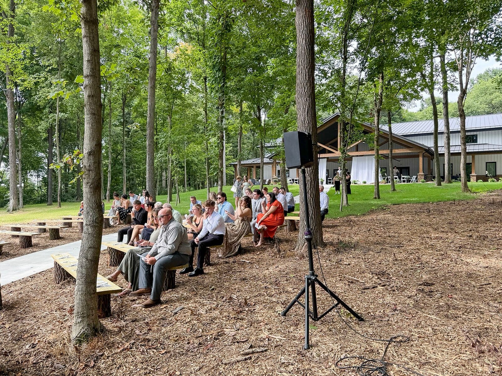 Guests seated among the trees for an outdoor ceremony at Highbanks Wedding & Events, with a ceremony speaker on a stand
